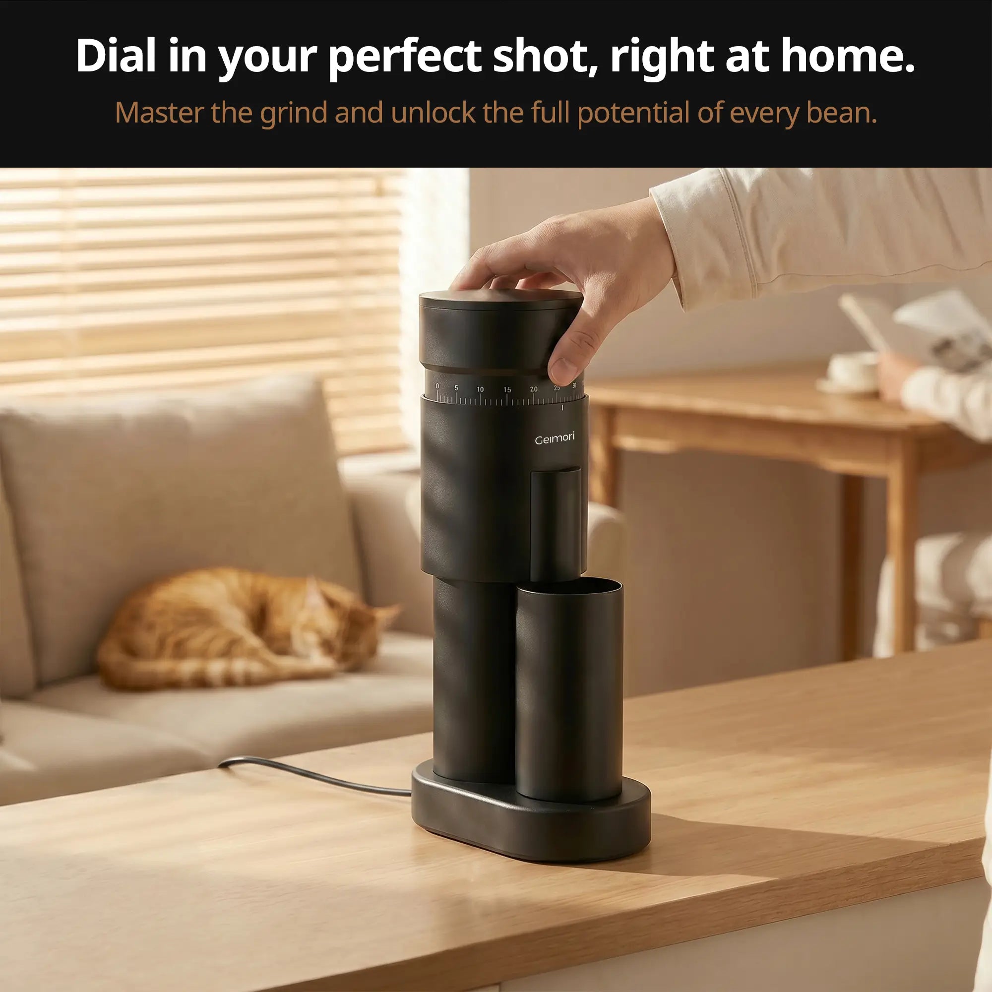 A black Geimori GU63 coffee grinder sits on a wooden countertop in a warm, modern living space. A hand is shown turning the top lid, with a cat sleeping on a beige sofa in the background.