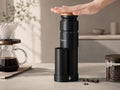 A black manual coffee grinder with a wooden top being pressed by a hand, set on a kitchen counter next to a glass pour-over coffee maker and a jar of coffee beans.