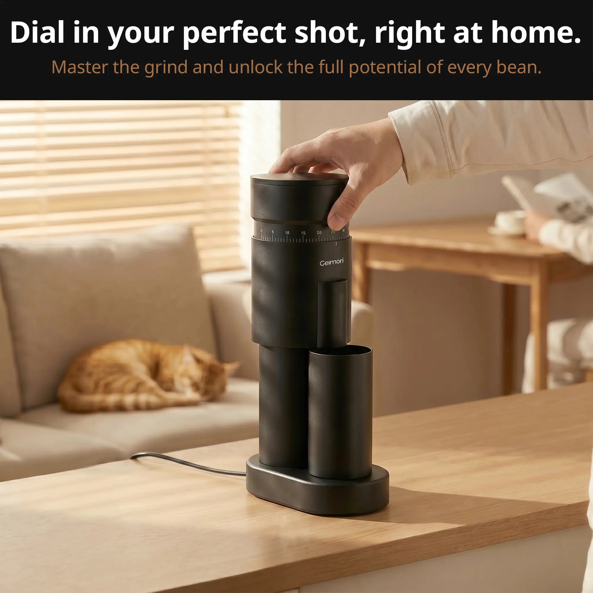 A black Geimori GU63 coffee grinder sits on a wooden countertop in a warm, modern living space. A hand is shown turning the top lid, with a cat sleeping on a beige sofa in the background.