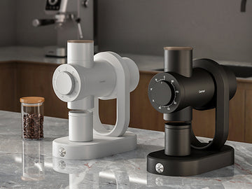 Two countertop coffee makers shown side by side on a marble-like kitchen counter, one in white and one in matte black, with a glass jar of coffee beans in the background.