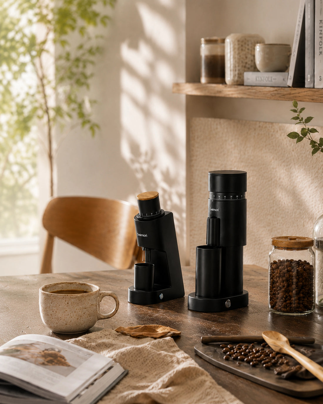 A black manual coffee grinder and bean hopper set on a wooden table in a warm, sunlit kitchen setting with a coffee cup, beans, and an open book nearby.slider_item_3FJgtJ