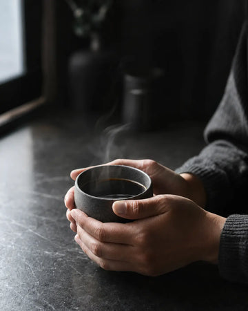 A person's hands holding a dark gray ceramic cup of hot coffee with visible steam rising, set on a dark tabletop in a dimly lit, moody environment.