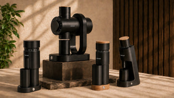 A set of black coffee brewing and serving equipment displayed on a tabletop, including a central coffee maker on a wooden base, two cylindrical brewers, and a bean hopper, styled against a warm beige wall and dark wood slats.